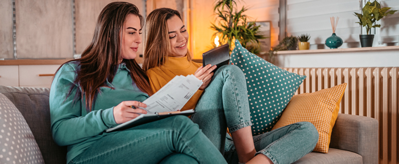 Two women sitting on a couch looking at paperwork and a calculator 