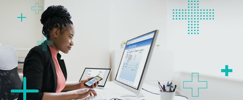 A woman holding a cell phone and looking at a computer screen