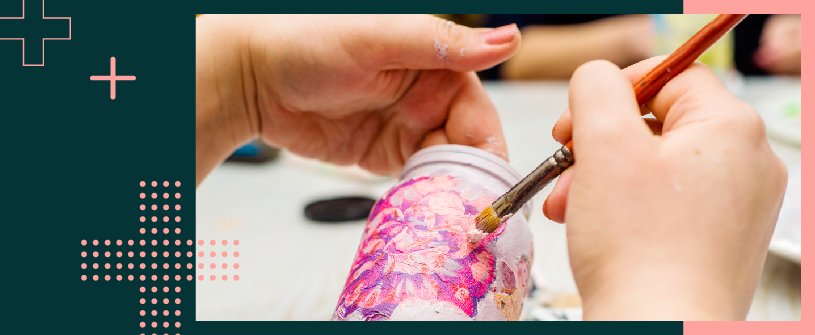 The hands of a person holding a brush and a jar, while they paint a flower on it. 