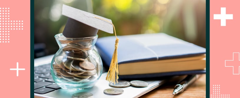 A jar filled with coins sitting overtop of a laptop with a graduation cap sitting overtop of it. A  book and a pen is laid beside it.